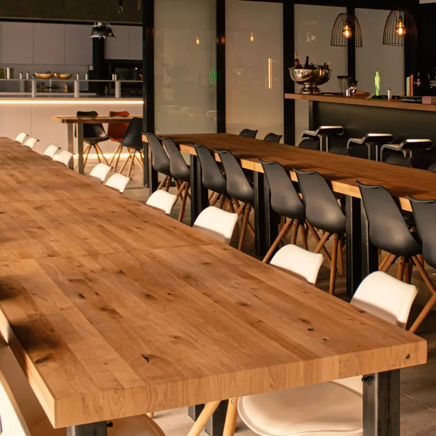 Mehrere lange Tischreihen mit Stühlen, im Hintergrund eine kleine Bar und ein Blick in eine Küche.